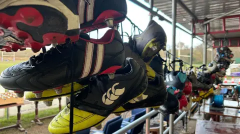 Close up shot of many pairs of rugby boots hanging on a line. There are a variety of colours although most of them are black boots with various sports brand logos on them. In the background there's a rugby pitch and some scaffolding poles are also visible.