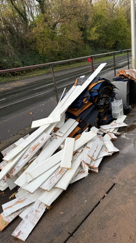 David Kyle Piles of white skirting boards and carpet piled up on the pavement next to a road. Dirt, debris and some water on the ground and in the background there are some trees with green and yellow leaves. 